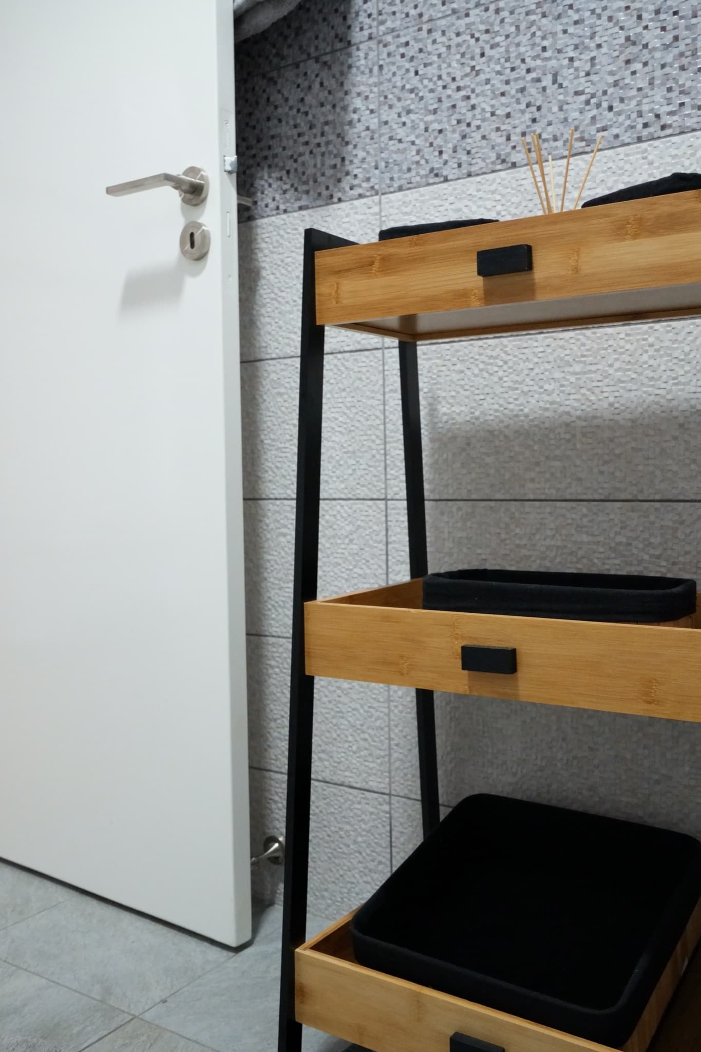Bamboo shelving unit with black trays beside white door and mosaic tiles