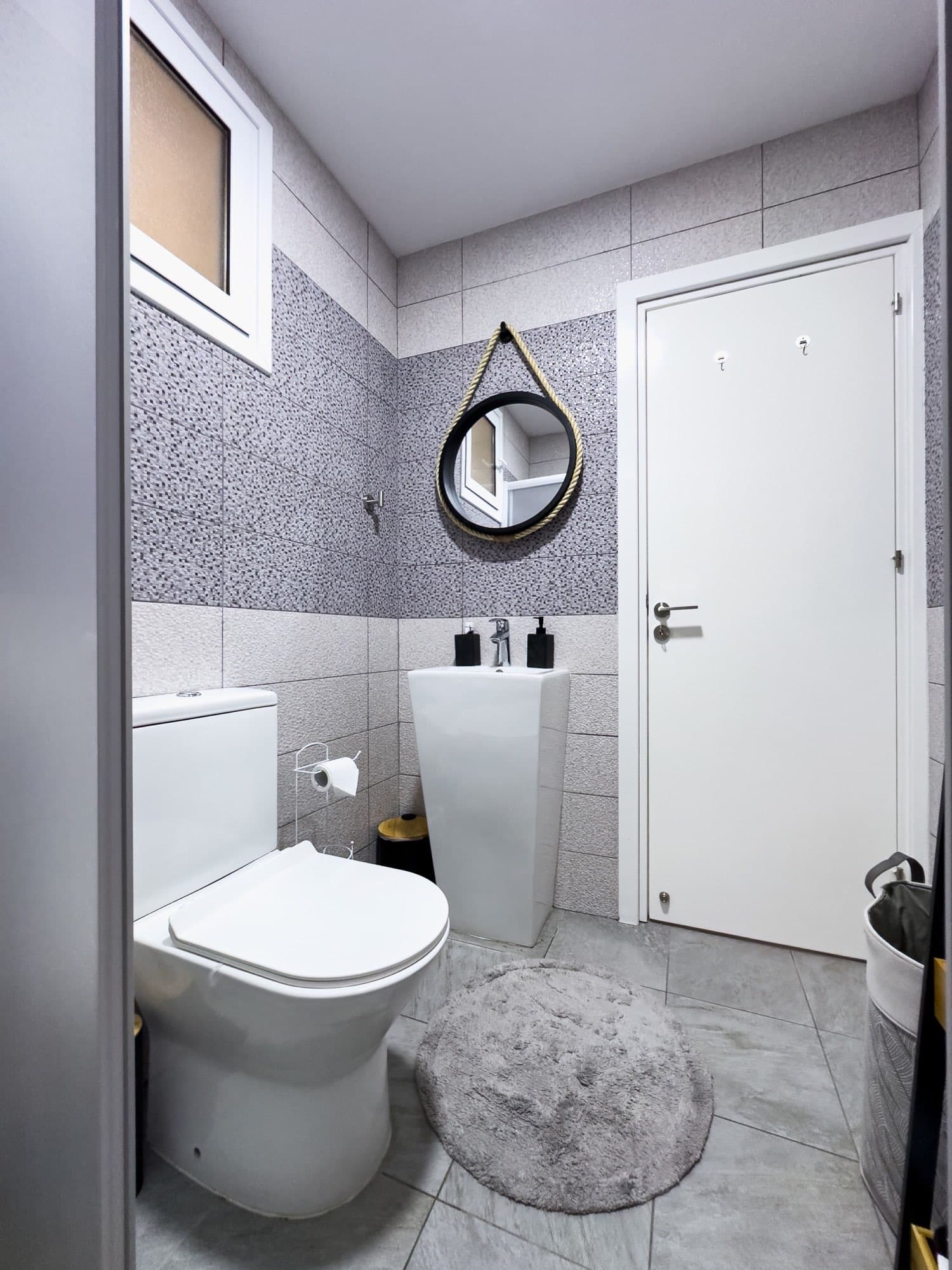 Bathroom with white toilet, pedestal sink, rope-framed round mirror, and mosaic tile walls