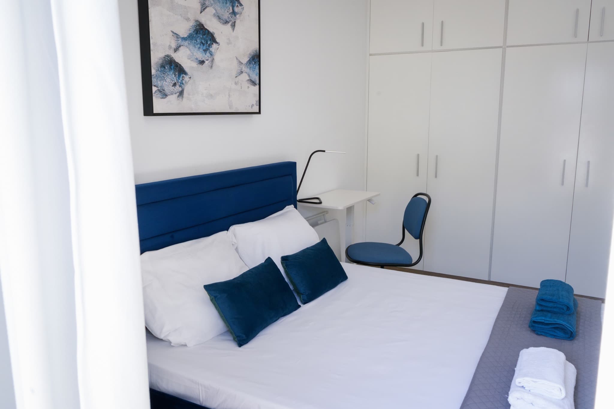 Blue upholstered bed with white pillows, towels, reading lamp, white wardrobes