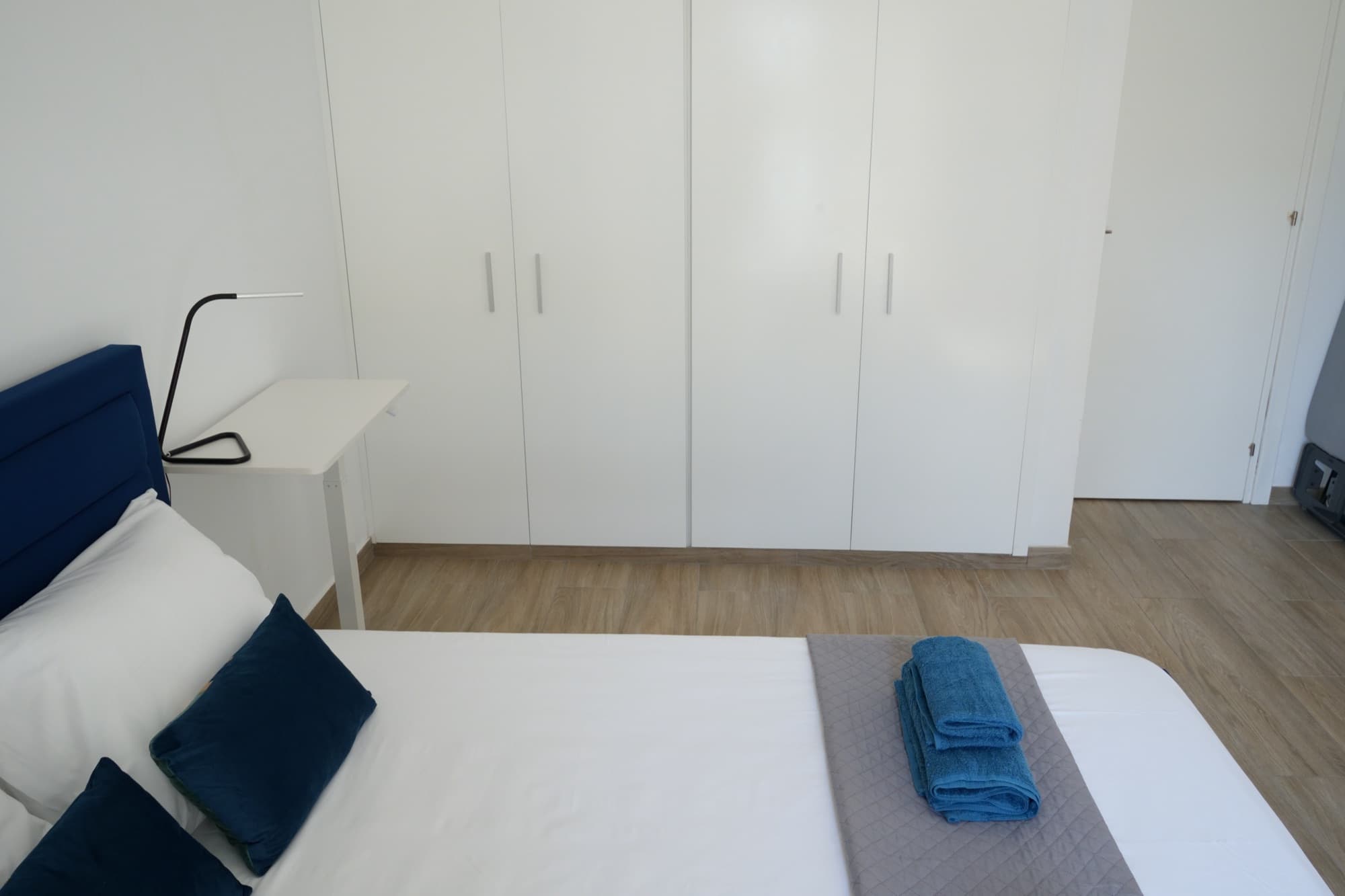 Blue bed with folded towels, white wardrobes, and bedside shelf