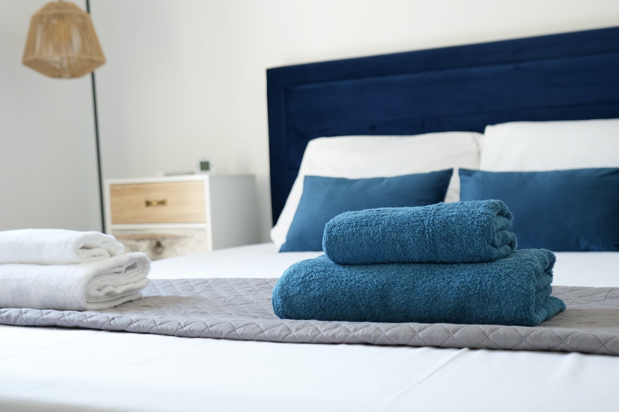 Blue bed with folded towels, wicker lamp, and two-drawer bedside cabinet
