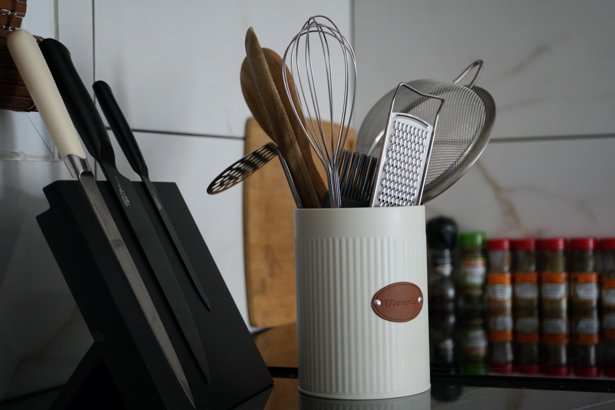 White ceramic utensil holder with whisk and spatulas beside knife block and spice rack