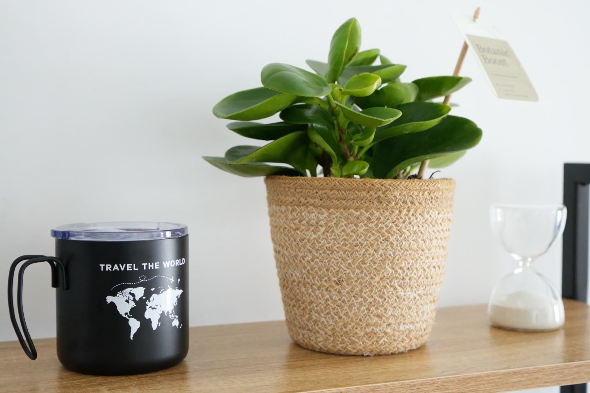 Black travel mug beside green plant in woven basket and glass hourglass on shelf