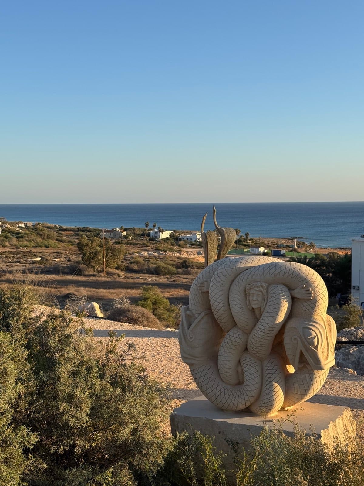 Stone serpent sculpture on plinth overlooking rocky coastline and Mediterranean sea