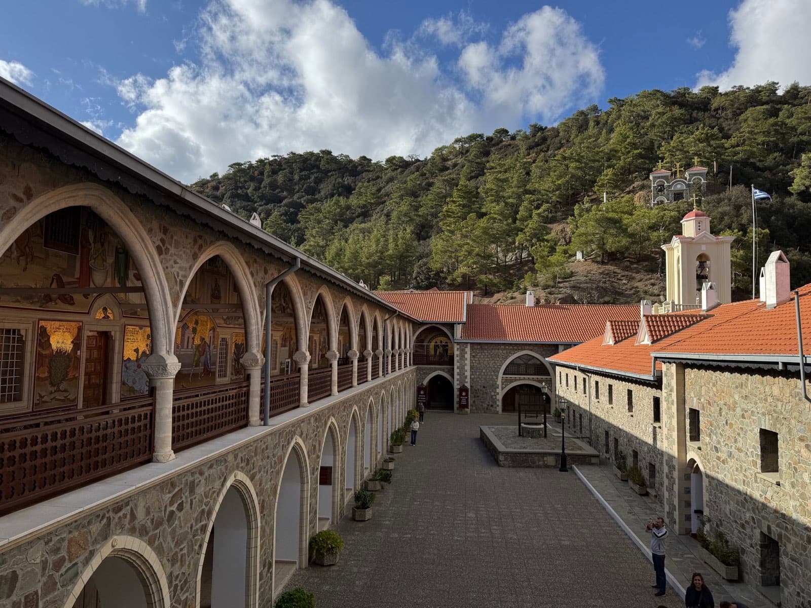 Kykkos Monastery courtyard with painted arched colonnade, bell tower, and forested hill