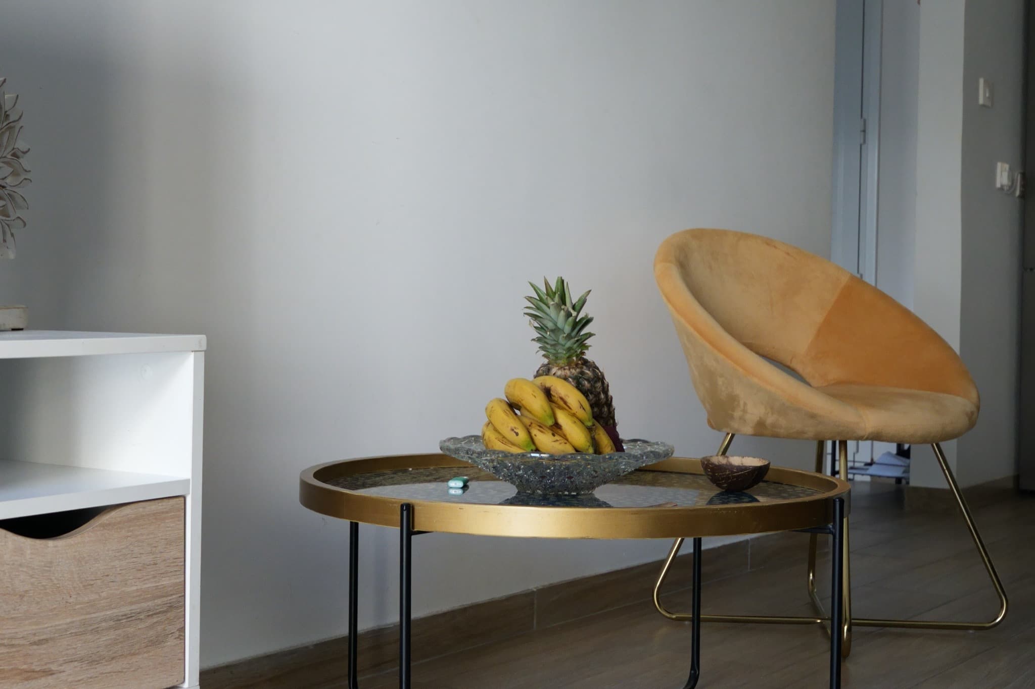 Round gold coffee table with bananas and pineapple beside yellow velvet scoop chair