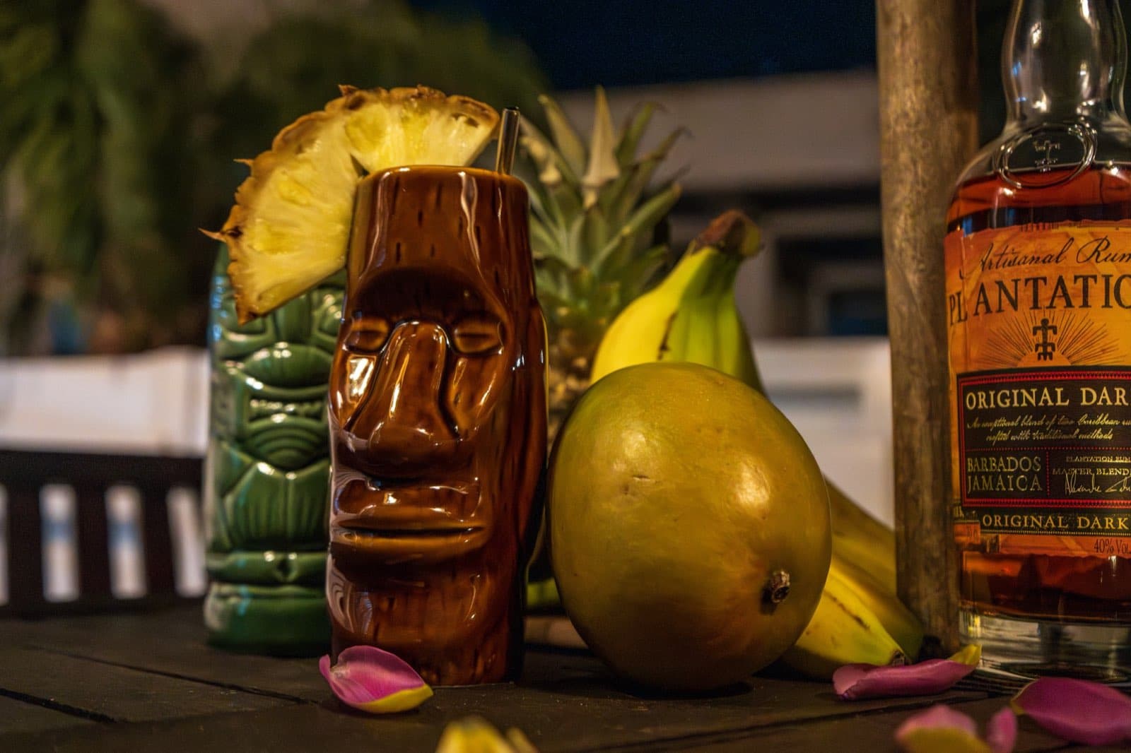 Brown tiki mug, green tiki mug, Plantation rum bottle, and tropical fruits on dark table