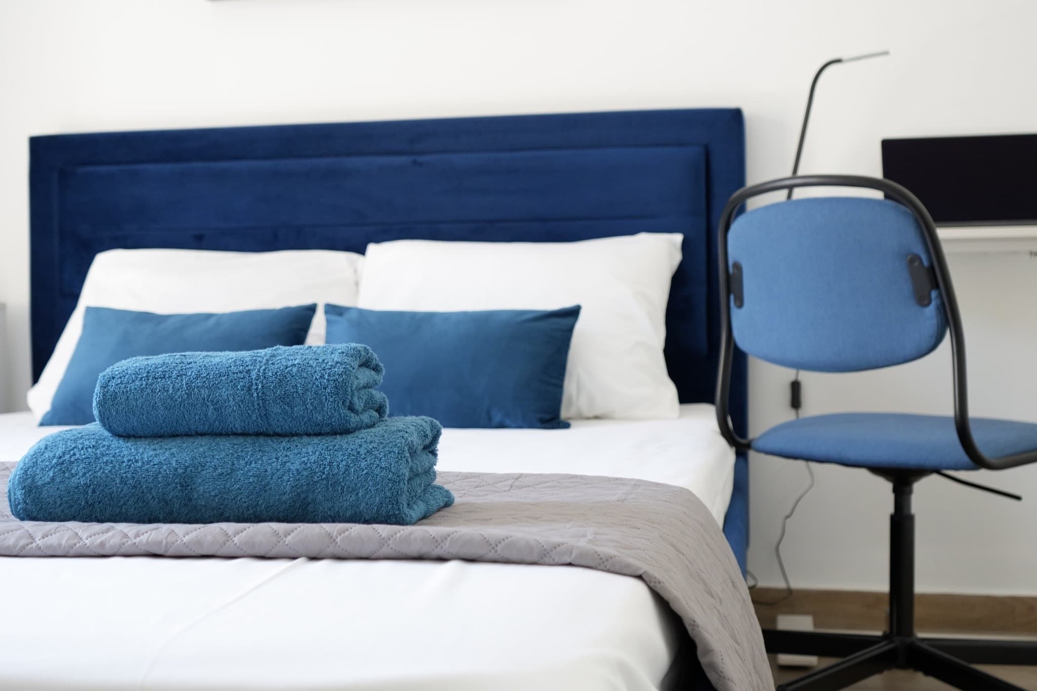 Blue upholstered bed with folded towels beside desk and blue chair