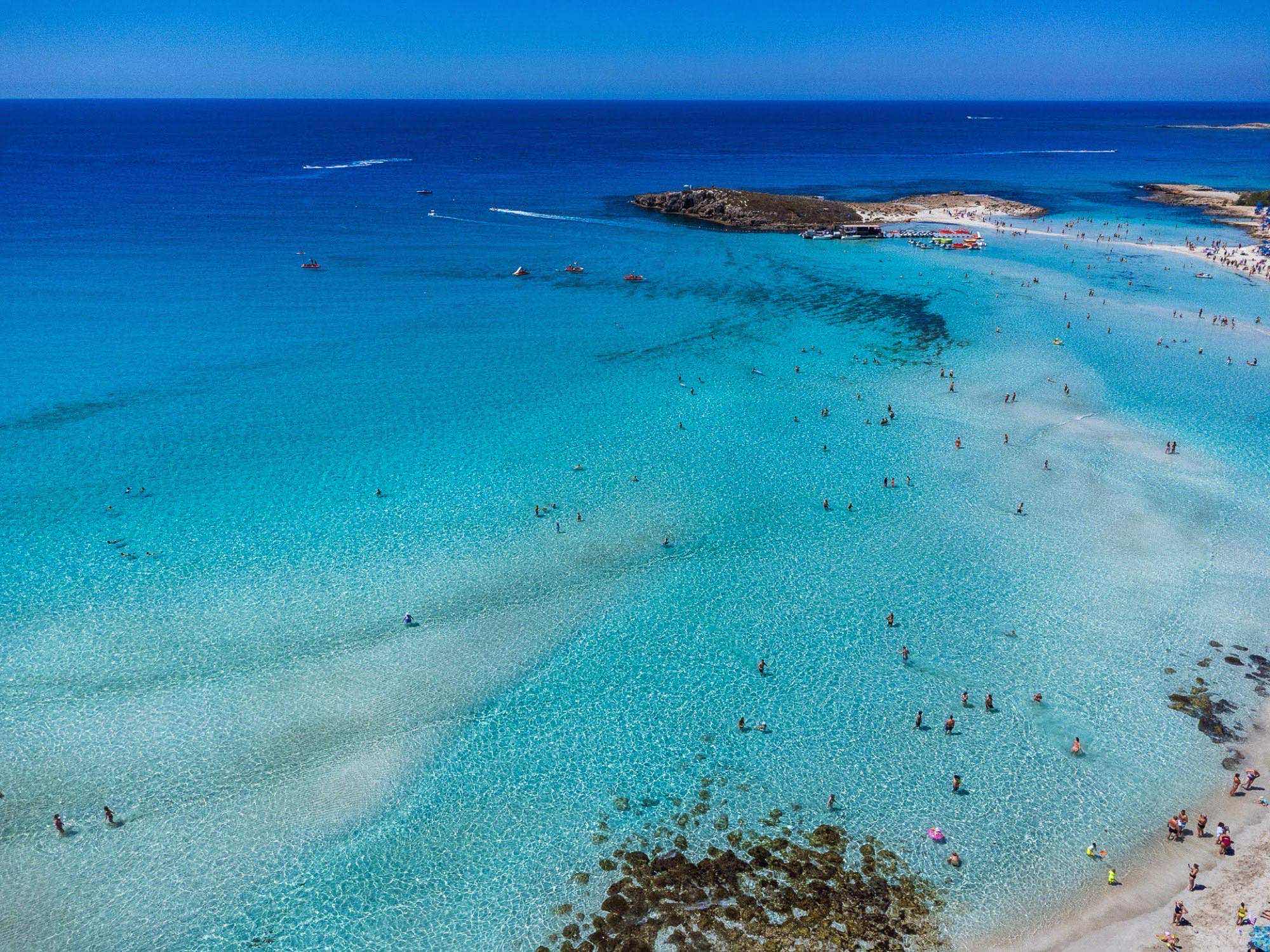 Aerial view of Nissi Beach's turquoise water and small islet, dotted with swimmers